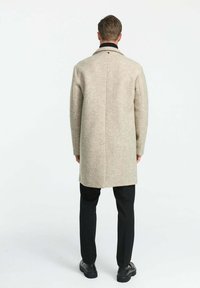Béžový vlnený kabát s rovnými líniami a zadným rozparkom, s čiernym golierom a jemnou textúrovanou vzorom. Nosený cez čierne nohavice a topánky.