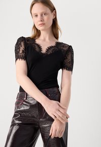 Camisole noire côtelée avec des manches en dentelle et un décolleté en forme de feston. Assortie à un pantalon sombre brillant orné de coutures rouges.