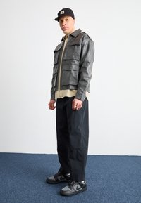 Siva usnjena jakna z več sprednjimi žepi, bež srajca spodaj, črne hlače in sivi Nike superge, postavljeni pred enostavno ozadje.