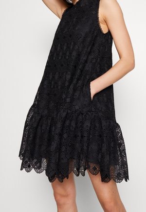 Czarna sukienka bez rękawów z koronką w kwiatowe wzory, z falowanym dołem i dłonią umieszczoną w bocznej kieszeni.