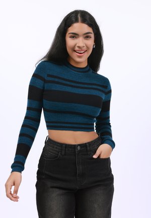 Pull cropped rayé en teal et noir, texture côtelée, col montant, associé à un jean noir taille haute, mains sur les hanches.