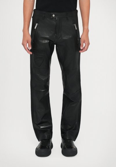 Pantalones de cuero negros con bolsillos con cremallera, logotipos en relieve de DSQUARED2 y Ducati en el muslo izquierdo, usados con zapatillas negras y una camiseta negra.
