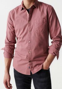Camicia di cotone rosa con bottoni, caratterizzata da piccoli pois scuri, taschino sul petto e maniche arrotolate. Indossata con jeans scuri.