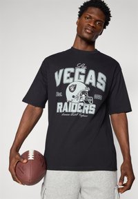 Juodas trumpomis rankovėmis marškinėliai su "Las Vegas Raiders" ir futbolo šalmą vaizduojančiu grafiniu elementu šviesiai pilkos spalvos. Modelis laiko rudą futbolą.