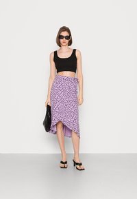Top crop preto, saia envolvente lavanda com padrão floral roxo, e sandálias de salto pretas. Modelo segurando uma bolsa texturizada preta.