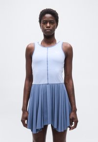 Γυναίκα που φοράει αμάνικη ανοιχτό μπλε μπλούζα και πλισέ μπλε φούστα μέτριου μήκους μπροστά σε απλό λευκό φόντο.