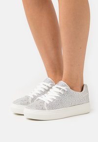 Silver glitter sneakers with white laces; textured surface detailed with sparkly embellishments; rounded toe and flat white sole.