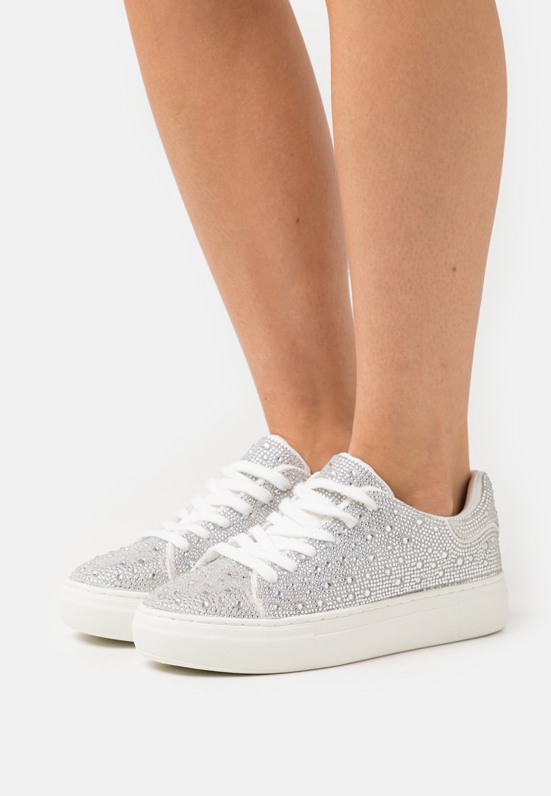 Silver glitter sneakers with white laces; textured surface detailed with sparkly embellishments; rounded toe and flat white sole.