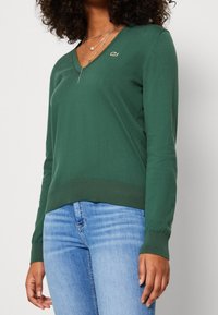 Grön V-ringad tröja i mjukt material med ribbad kant och en liten logodetalj på bröstet. Matchas med blå jeans.