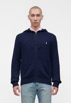 Homme à la tête rasée portant un sweat à capuche zippé bleu marine en maille torsadée et un jean bleu clair, debout devant un fond clair uni.