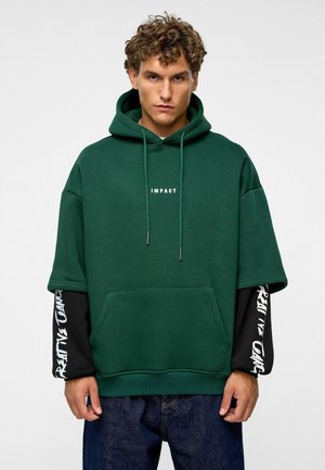 Groene oversized hoodie met voorzak, trekkoord capuchon en witte tekst "IMPACT". Zwarte lange mouwen met witte tekstpatronen. Denim jeans.
