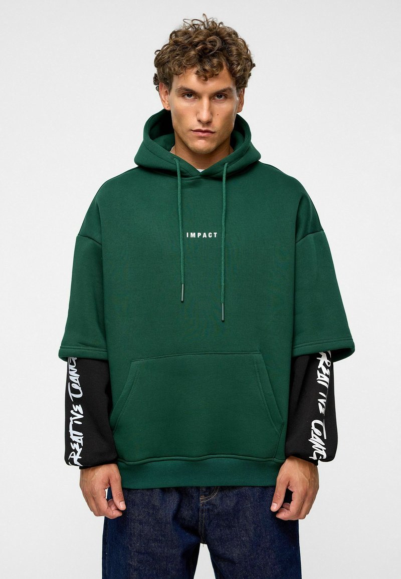 Haut à capuche oversize vert avec poche avant, capuche à cordon, et texte blanc "IMPACT". Manches longues noires avec des motifs de texte blancs. Jean en denim.