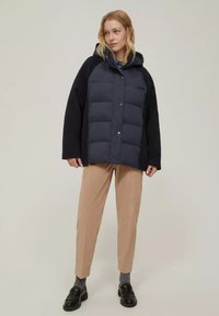 Giacca a vento blu navy con cappuccio e motivo a rombi, con maniche nere. Abbinata a pantaloni beige e mocassini neri.