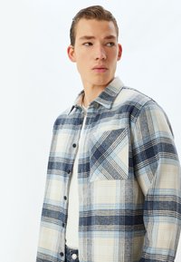 Camisa de franela ligera en cuadros de azul, navy y crema, con un bolsillo en el pecho y cierre de botones, con una textura suave y diseño clásico.