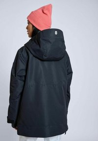 Svart vattentät jacka med huva, med en avslappnad passform och textilstruktur. En rosa mössa sitter på bärarens huvud.