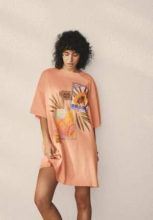 Frau mit lockigem Haar, die ein übergroßes pfirsichfarbenes Kleid mit tropischen und Brasilien-Motiven trägt, steht vor einer strukturierten, cremeweißen Wand.