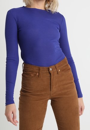 Personne portant un haut bleu moulant à manches longues et un pantalon en velours côtelé marron taille haute, debout devant un fond clair et uni.