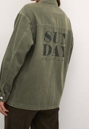 Person trägt eine olivgrüne, übergroße Jacke mit dem schwarzen Aufdruck "SUNDAY Chill out" auf dem Rücken, kombiniert mit dunkelbraunen Hosen.
