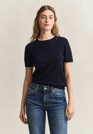 Jeune femme aux cheveux ondulés portant un haut bleu marine à manches courtes et un jean bleu, debout devant un fond beige uni.