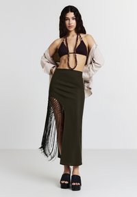 Gonna verde oliva con una alta spacco e frange nere, abbinata a un top bikini marrone e un cappotto beige. Scarpe con piattaforma nere completano il look.