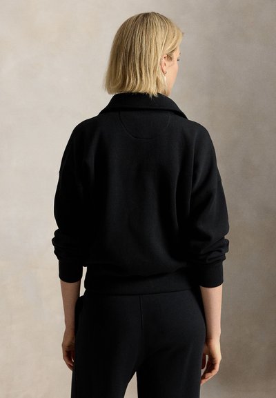 Donna bionda vista da dietro che indossa una maglia nera a maniche lunghe e pantaloni neri su uno sfondo beige uniforme.