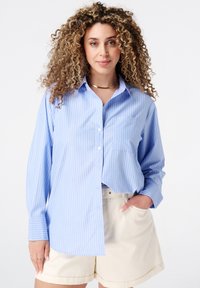 Chemise boutonnière bleu clair avec des rayures verticales blanches, manches longues, et une poche sur le côté gauche, associée à un short beige.