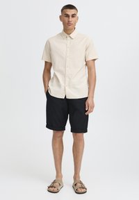Beiges kurzärmliges Hemd mit Knöpfen, schwarze hochgekrempelte Shorts und braune Sandalen. Das Outfit besticht durch ein schlichtes, sauberes Design mit lockerer Passform.