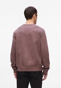 Sudadera de algodón marrón con cuello redondo y puños de canalé, que presenta un logo bordado en la parte inferior, combinada con pantalones negros.