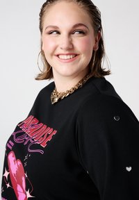 Zwarte sweatshrit met gedurfde roze tekst en hartgrafieken. Zilveren hartaccenten op de mouwen. Glad textuur, ontwerp met ontspannen pasvorm.