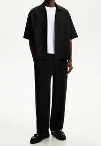 Czarna koszula z krótkim rękawem i kołnierzykiem, noszona na białej koszulce, zestawiona z luźnymi czarnymi spodniami i czarnymi butami. Czysty, minimalistyczny design.