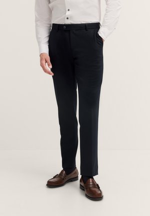 Mann trägt schwarze Anzughose, weißes Hemd mit Knöpfen und braune Lederschuhe, steht mit einer Hand in der Hosentasche vor einfarbigem Hintergrund.