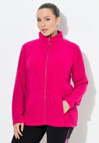 Kvinna med håret i en knut, iklädd en ljusrosa zip-up fleecejacka, som står mot en vit vägg med en hand i fickan.