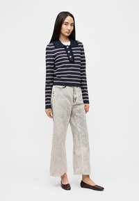 Μπλούζα με μακριά μανίκια σε ναυτικό ριγέ σχέδιο, με γιακά και τρία κουμπιά, συνδυασμένη με ανοιχτό γκρι τζιν παντελόνι φαρδιάς γραμμής και σκούρα καφέ επίπεδα παπούτσια. Ομαλές υφές εμφανείς.