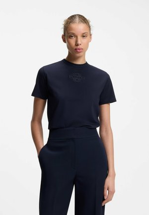 Frau mit geflochtenem Haar trägt ein marineblaues Kurzarmshirt und passende Hose, steht mit einer Hand in der Tasche vor einem weißen Hintergrund.