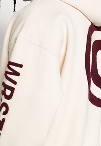 Krémová mikina s kapucí z měkké látky, s vínově červeně vyšívanými logy na hrudi a rukávu. Mírně oversize střih.