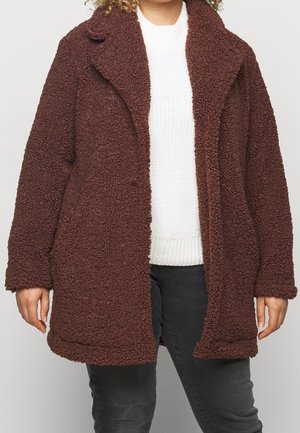 Manteau en laine brun texturé porté sur un pull blanc en maille et un pantalon gris foncé, montré sur une personne du cou jusqu'au milieu de la cuisse.