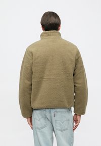 Olívazöld polárdzseki magas gallérral, laza szabással és texturált felülettel. Látható varrásokkal és enyhén túlméretezett ujjak, világoskék farmerral párosítva.