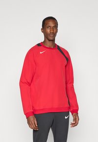 Czerwona sportowa bluza z okrągłym dekoltem, czarnymi akcentami na ramionach i logo Nike. Gładka tekstura i luźny krój. Połączona z ciemnymi spodniami.