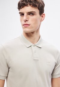 Ανοιχτό μπεζ πόλο πουκάμισο με κλασικό γιακά, δύο κουμπιά και υφασμάτινη υφή. Διαθέτει μικρό ανάγλυφο λογότυπο στο αριστερό στήθος.