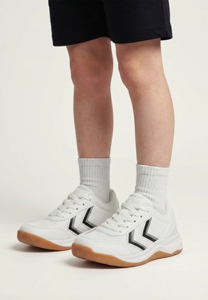 Kind draagt witte sneakers met zwarte chevronstrepen en gumzolen, gecombineerd met witte sportsokken en zwarte shorts.