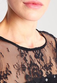 Svart spetsblus med blommönster, rundad halsringning och transparent material. Utsmyckad med små dekorationer nära kragen.