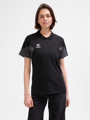 Vrouw met kort bruin haar die een zwart en donkergrijs poloshirt met hummel-logo draagt, staand tegen een effen lichte achtergrond.