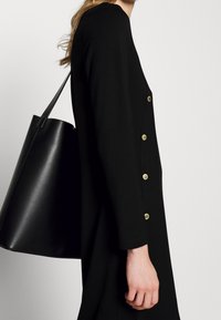Černé tunikové šaty s dlouhými rukávy, vyznačující se zlatými knoflíkovými akcenty na bočním okraji. Na jedné straně je držena černá kožená taška.