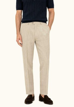 Homme portant un pantalon habillé beige, des mocassins marron foncé et une chemise en maille bleu marine à manches courtes, debout les mains dans les poches.