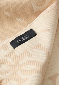 Beige kuvioitu kangas, jossa on poimutettu rakenne ja musta "GUESS"-etiketti ommeltuna kulmaan. Mallissa on hienovaraisia kuvioita.