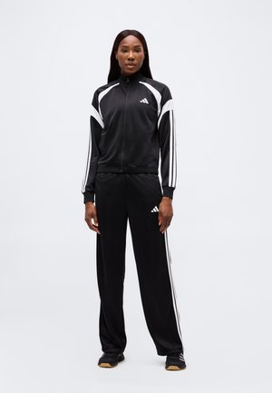 Mujer de pie vistiendo un chándal Adidas en blanco y negro con chaqueta con cremallera y pantalones a juego contra un fondo blanco liso.
