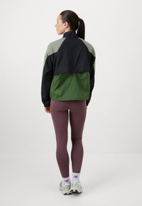 Veste coupe-vent colorée en noir, olive et vert. Matériau léger avec un design court et un col haut. Associée à des leggings bordeaux.