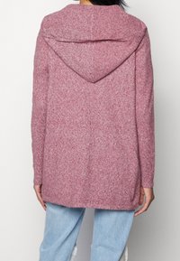 Lange, gezellige roze trui met een zachte textuur, capuchonontwerp en zijsplitten, gecombineerd met lichtblauwe spijkerbroek.