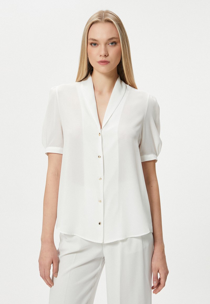 Blusa bianca a maniche corte con colletto a scialle, caratterizzata da una chiusura frontale con bottoni e una leggera lucentezza, realizzata in tessuto leggero e traslucido.