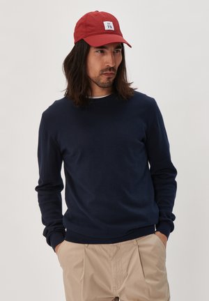 Marineblauwe gebreide trui met geribbelde manchetten en zoom, gecombineerd met een rode pet met een wit label. Model draagt beige broeken.